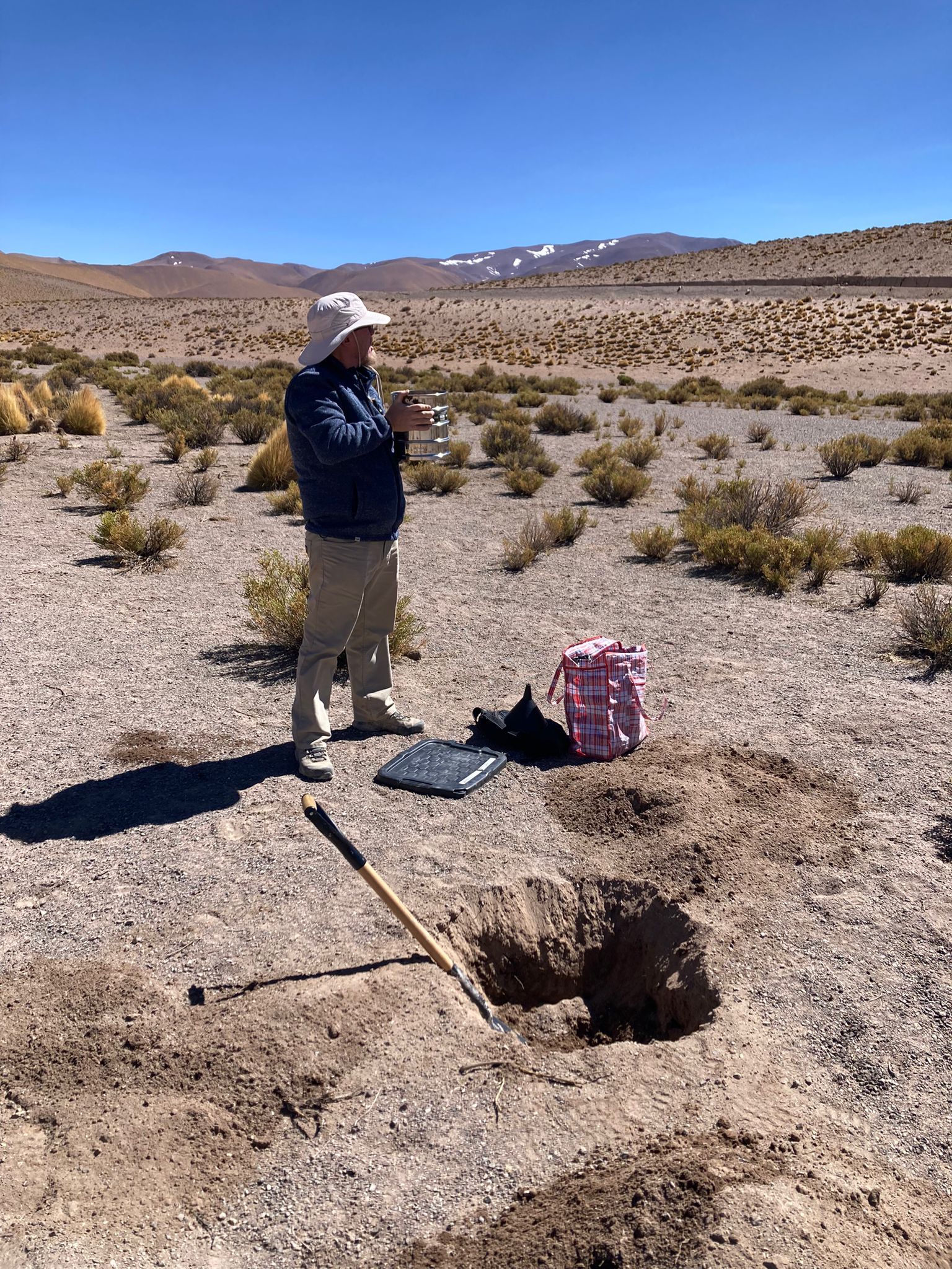 Geologist with a stack of sieves beside a freshly dug soil sample pit and shovel