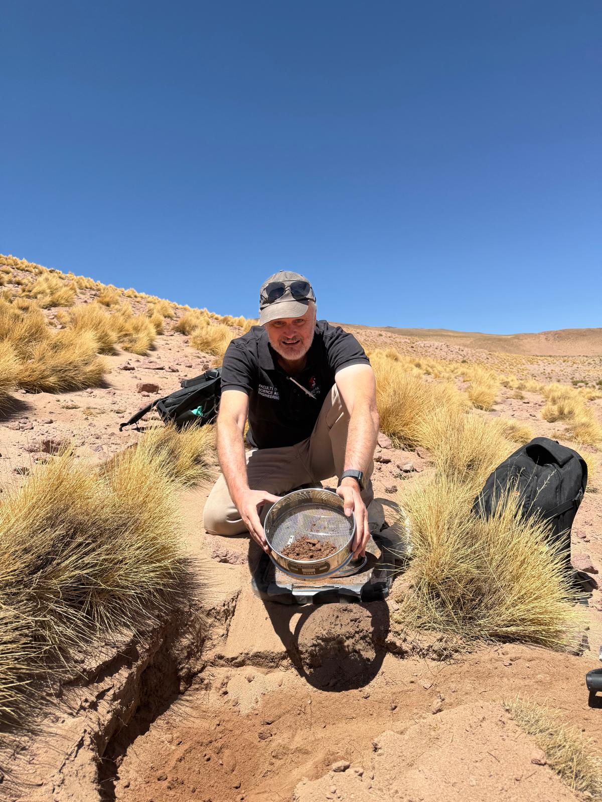 Geologist kneeling beside a pit holding a sieve of fine soil sample