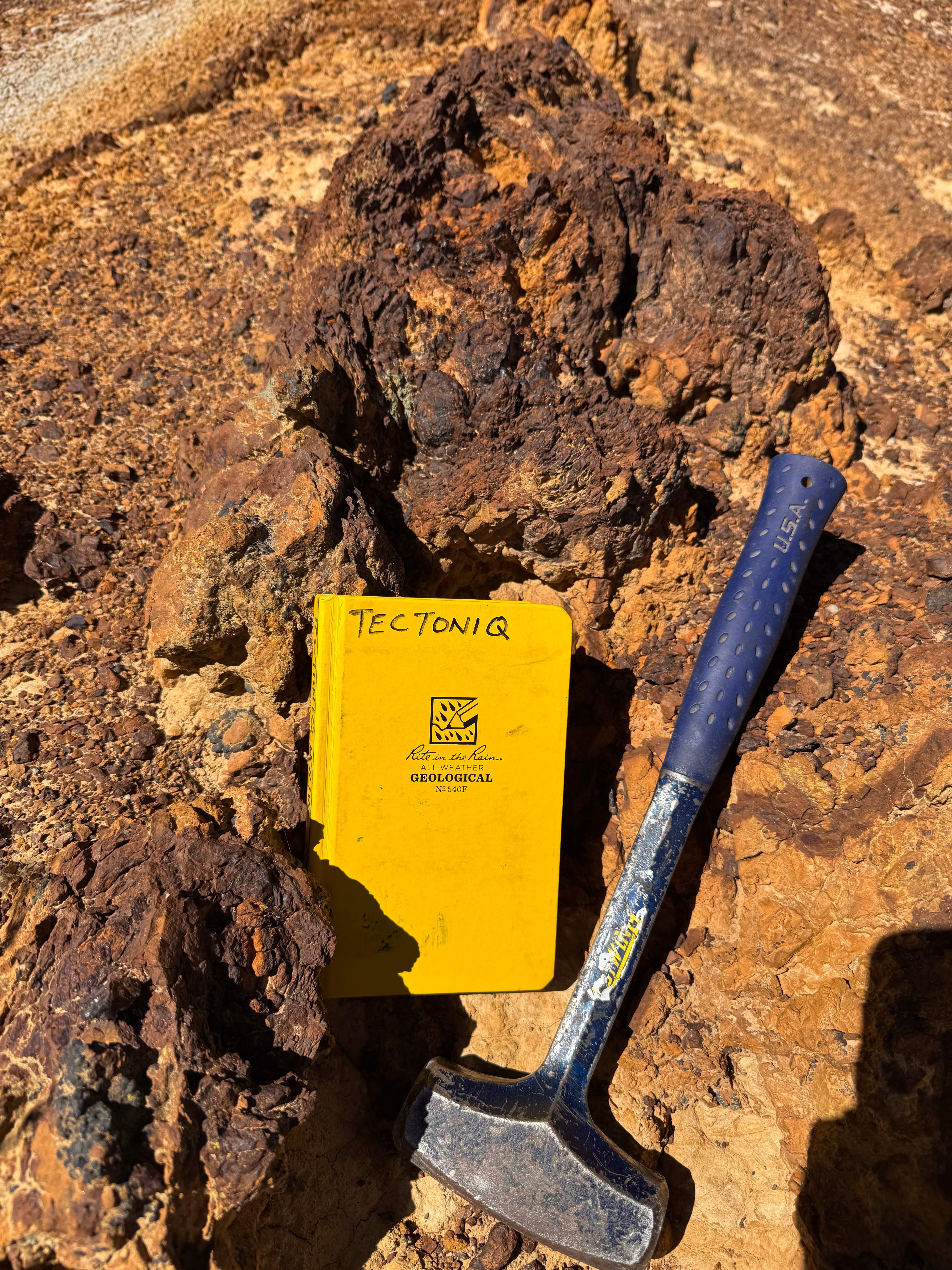 Tectoniq field notebook and geological hammer resting on an iron-stained gossan outcrop
